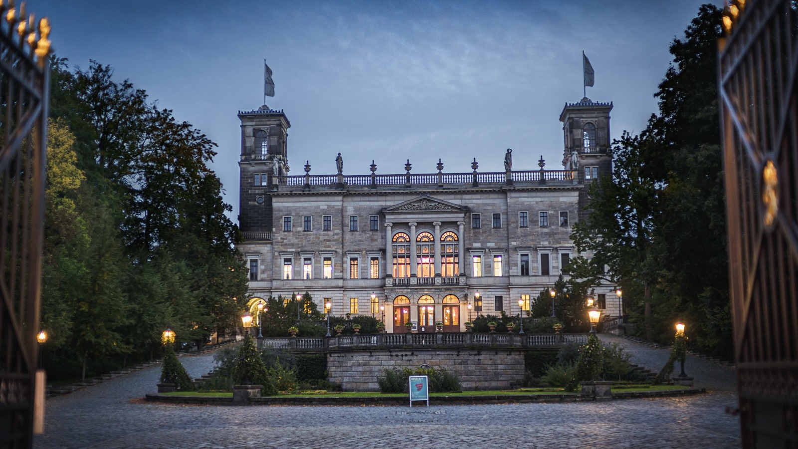 Beleuchtetes Palais in der Dämmerung hinter einem schmiedeeisernen Tor